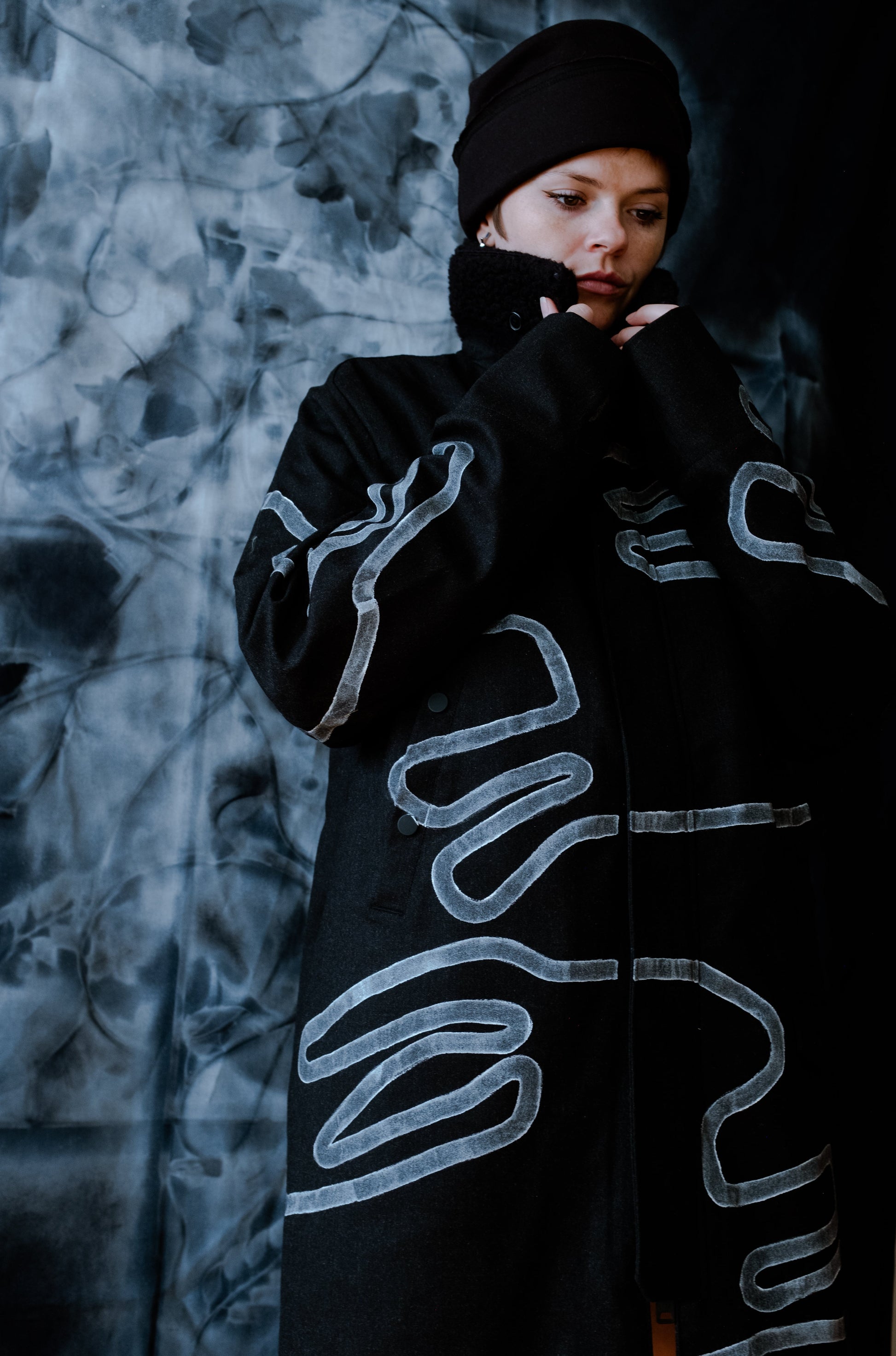 Model standing in front of cloudy blue backdrop wearing black long winter coat with faux shearling collar and white hand painted rabbit silhouette designs across the front and arms