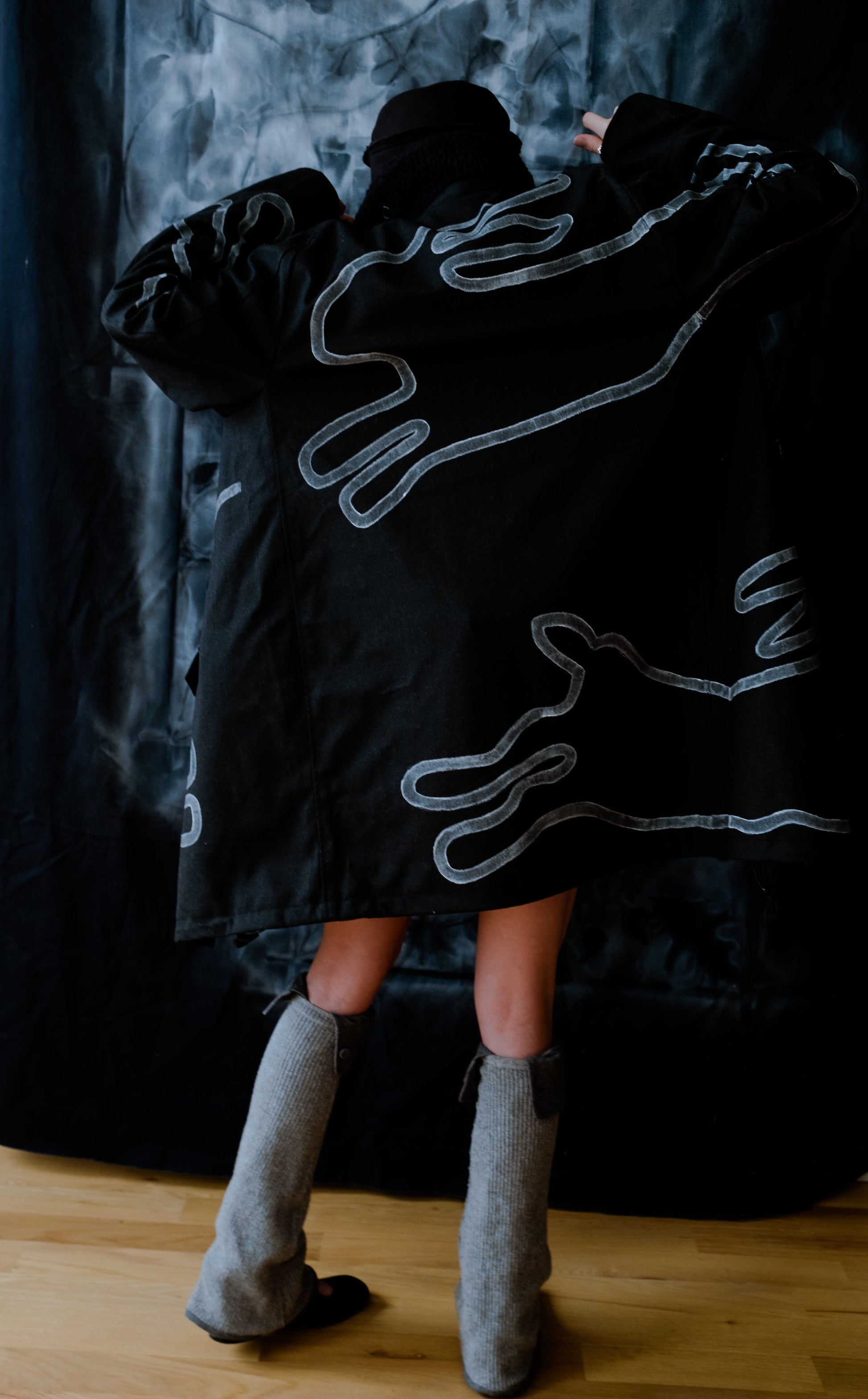 back of model wearing black long winter coat with faux shearling collar and white hand painted rabbit silhouette designs across the back and arms