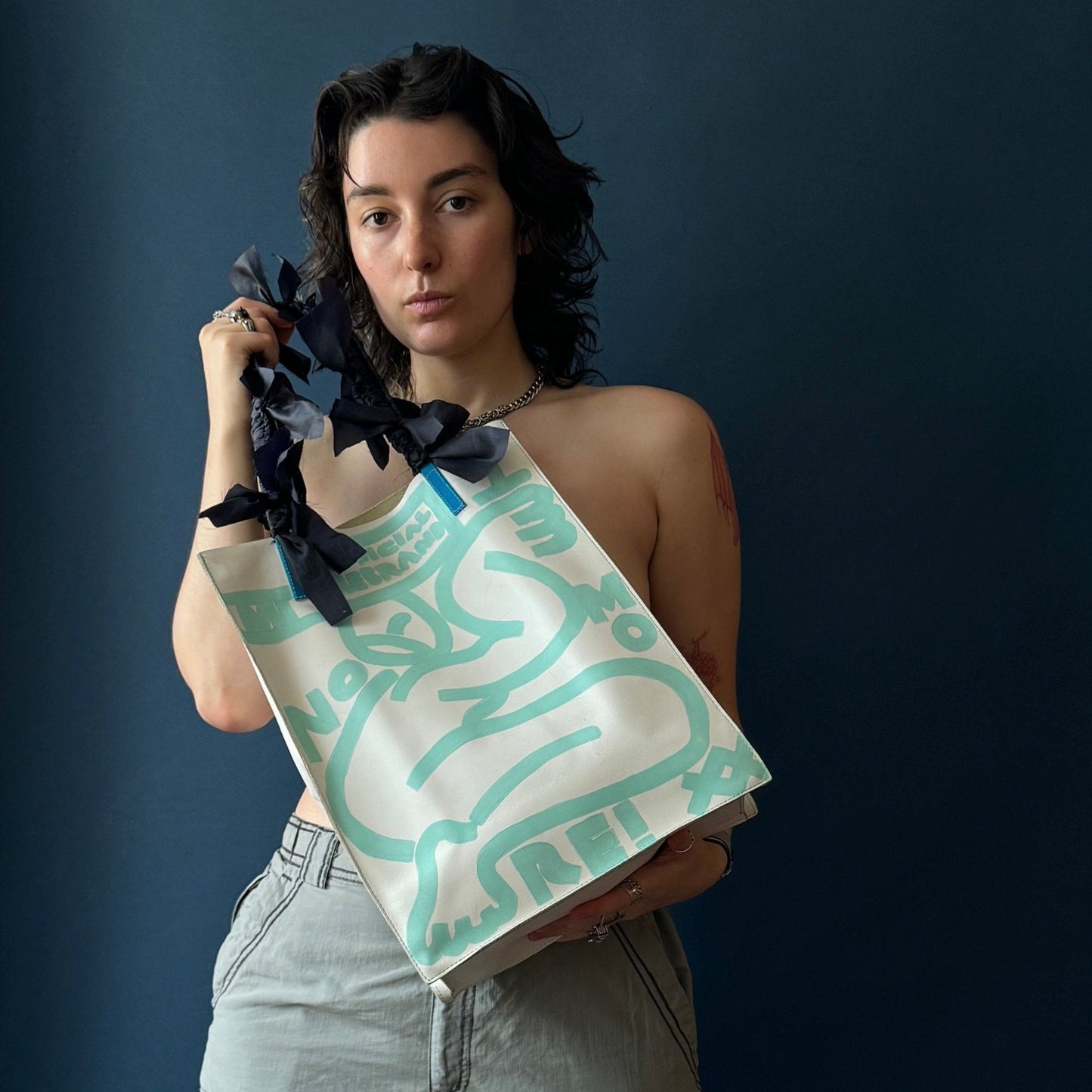model holding a rectangular white leather bag with aqua painting of a figure and text reading NO MORE. Bag handles have navy fringe