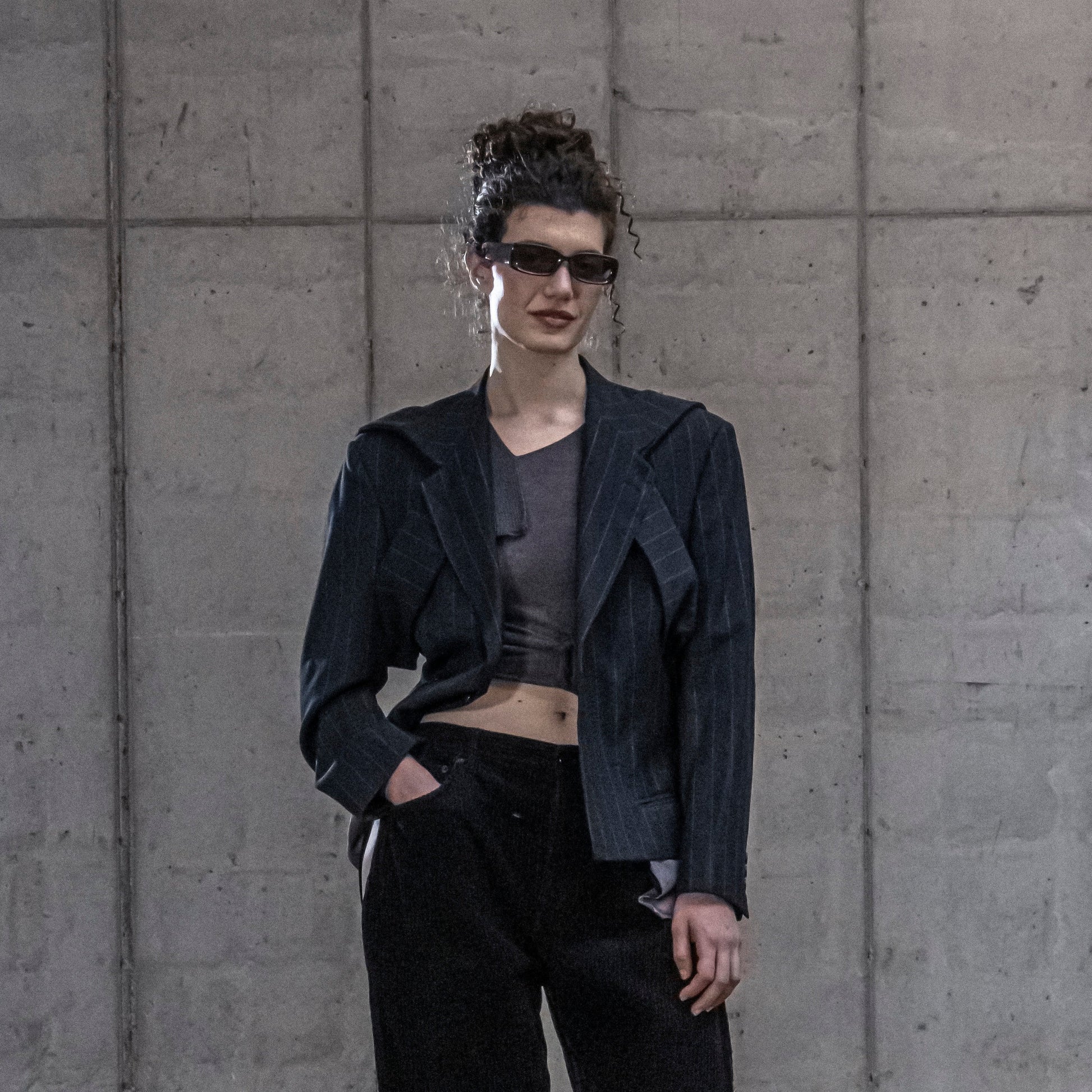 Person wearing a pinstripe jacket and sunglasses against a gray concrete wall