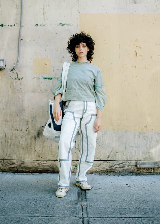 A person wearing a long-sleeved, light green T-shirt with extended sleeves and a round neckline, paired with white pants featuring a striped pattern. The person is standing against a neutral background.