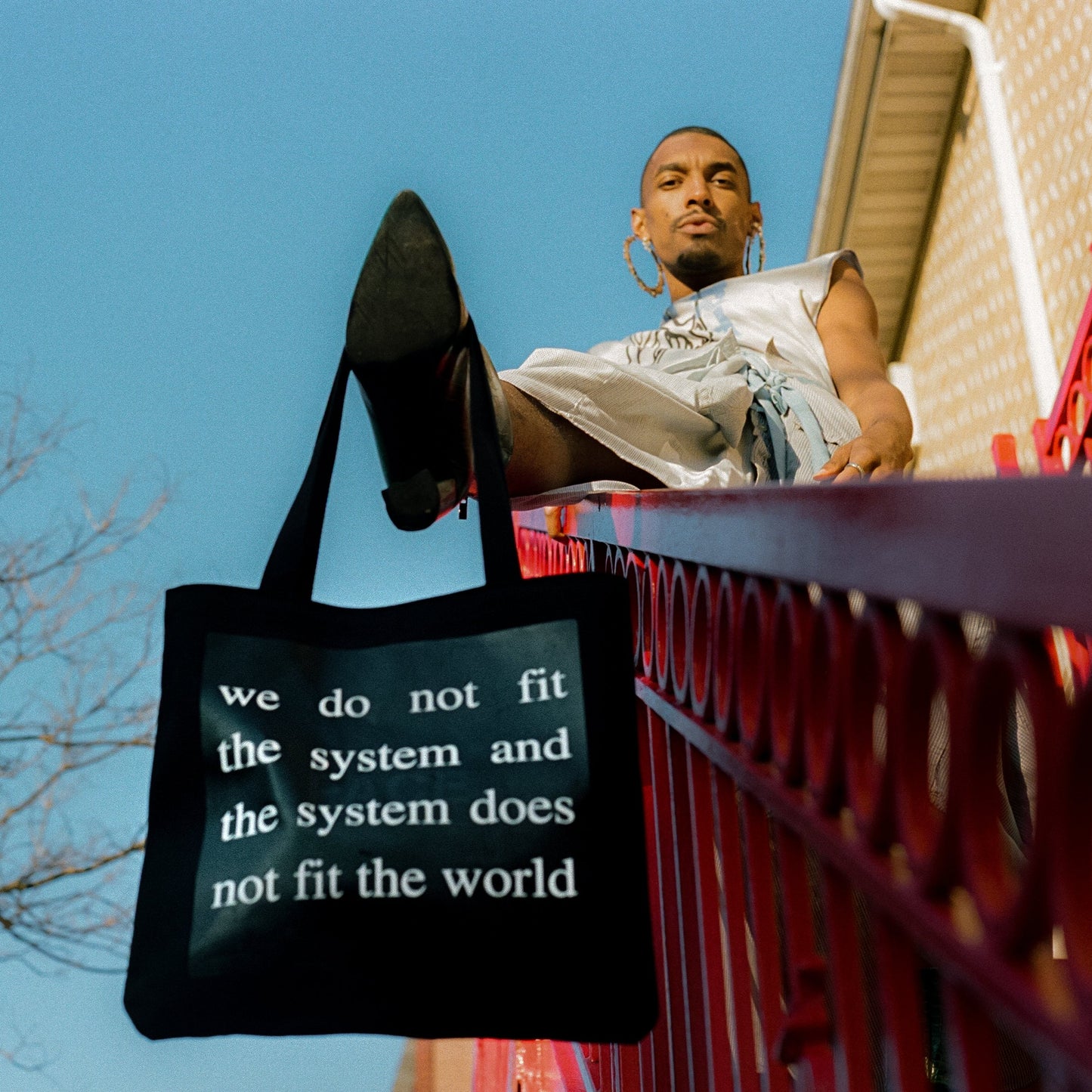 model using their foot to hold black tote bag with white screen printed text reading we do not fit the system and the system does not fit the world
