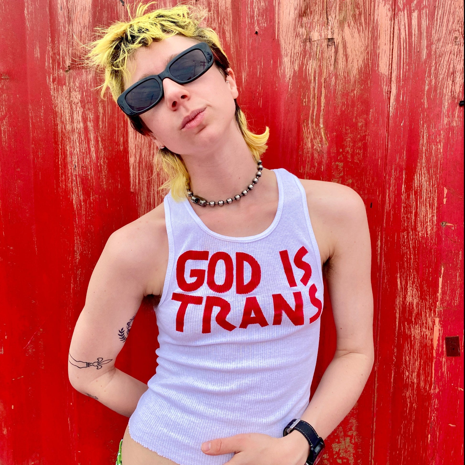 model wearing white ribbed A-shirt tank top with hand painted text reading GOD IS TRANS in red across the chest