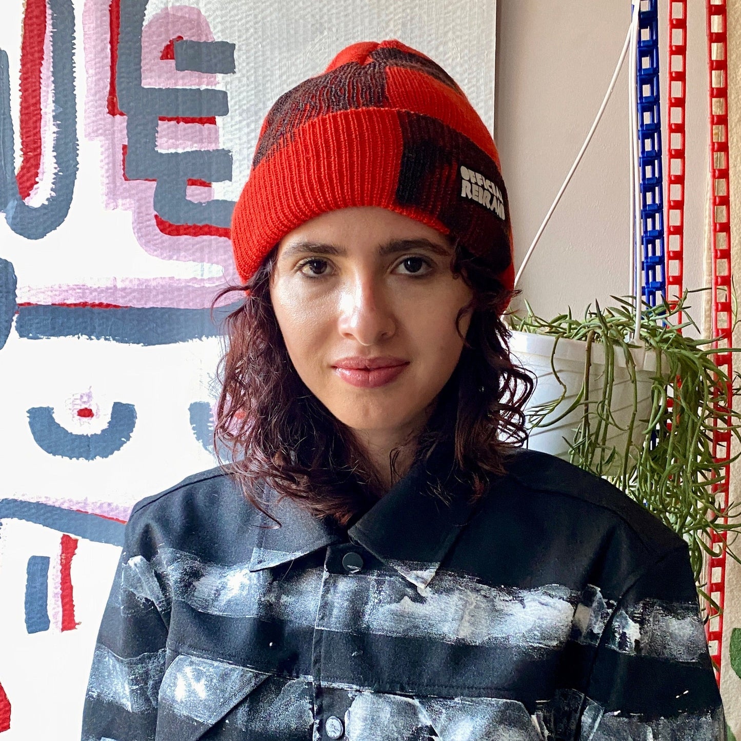 Model wearing red beanie with hand painted black checkered squares and a black tag with white text reading OFFICIAL REBRAND on the hat's cuff