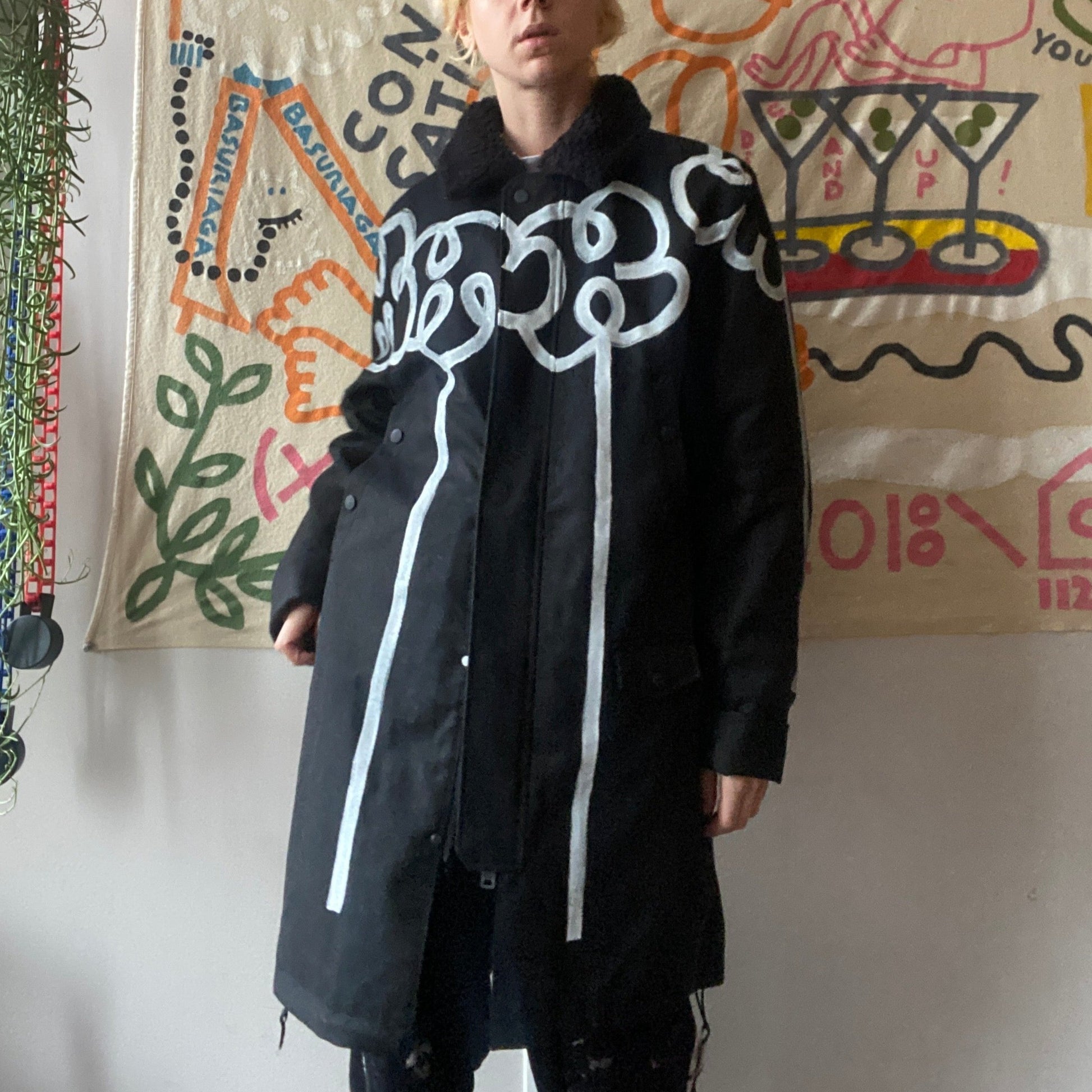 Model wearing black long winter coat with faux shearling collar and white hand painted flower designs across the front and arms