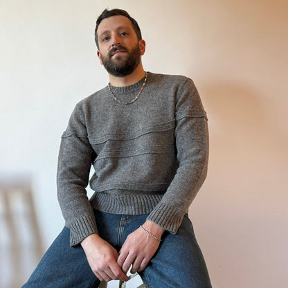 Man wearing a gray sweater and blue jeans sitting on a chair against a plain wall.