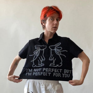 Model wearing cropped black collared polo shirt with two white hand painted rabbits facing each other and white text reading I'M NOT PERFECT BUT I'M PERFECT FOR YOU