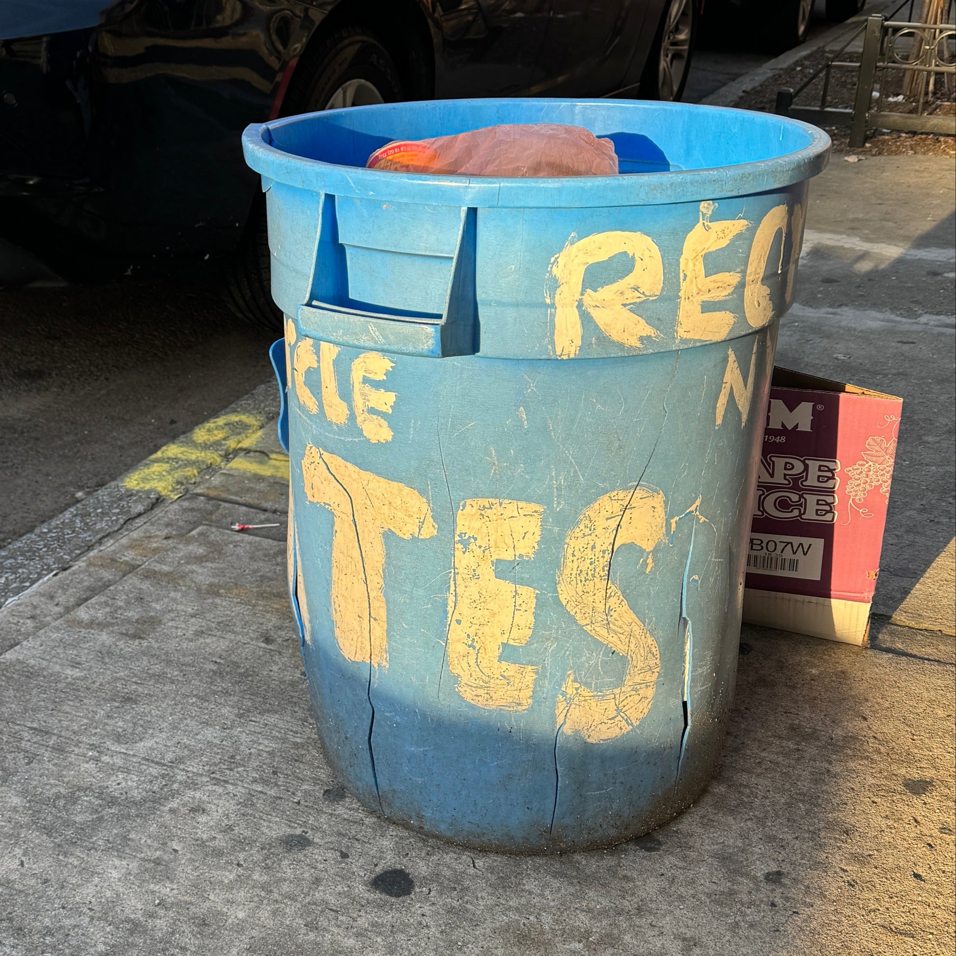 Blue recycling bin with text on a sidewalk next to parked cars