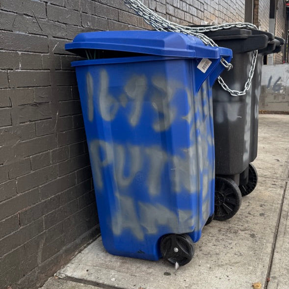 Blue trash bin with graffiti on a sidewalk next to a building.
