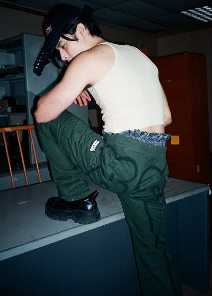 Person in a white tank top and green pants standing on a counter in a room with a wooden chair and shelves.