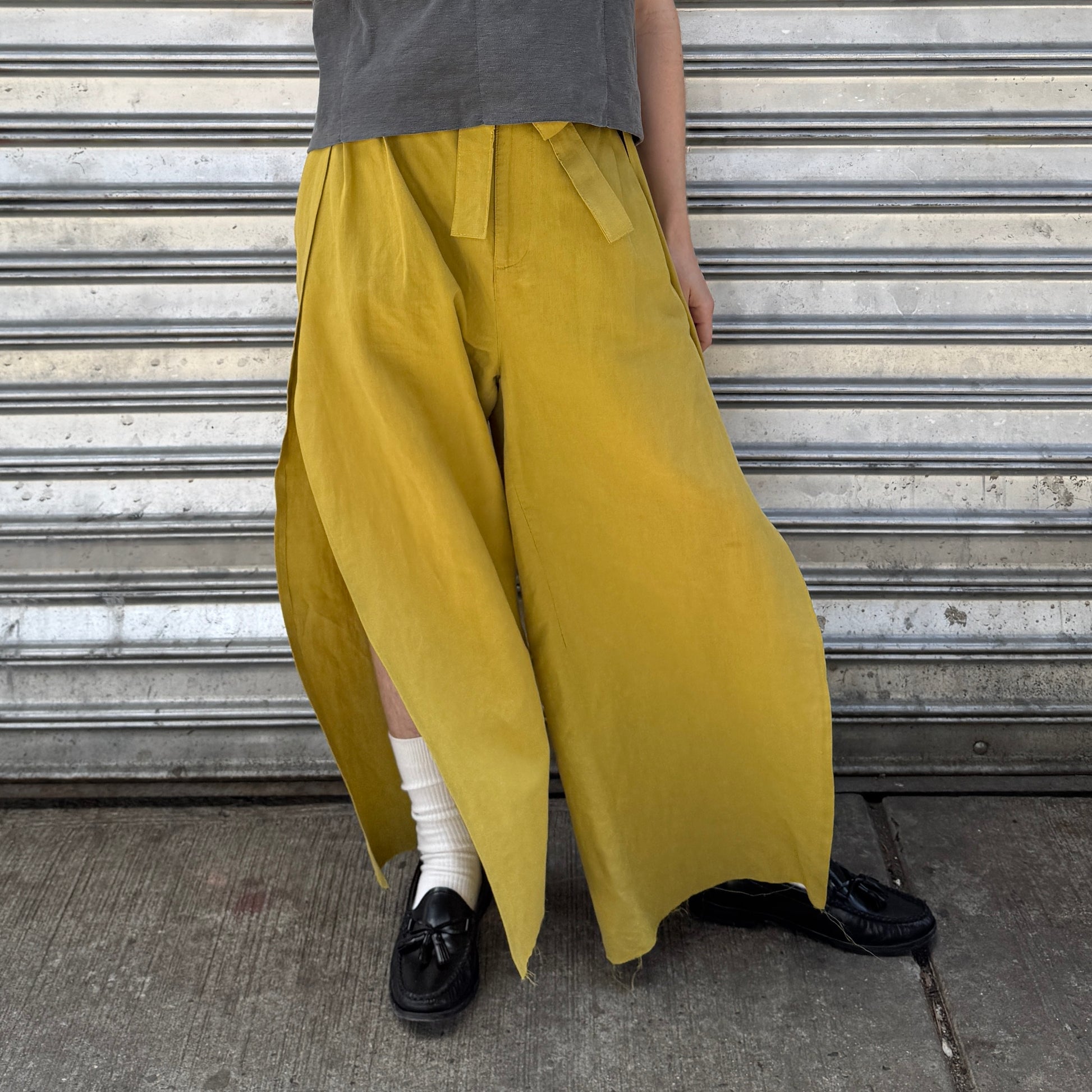 Person wearing mustard yellow wide-leg pants with a gray shirt and black shoes against a metal shutter background.