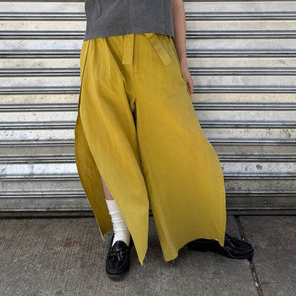 Person wearing mustard yellow wide-leg pants with a gray shirt and black shoes against a metal shutter background.