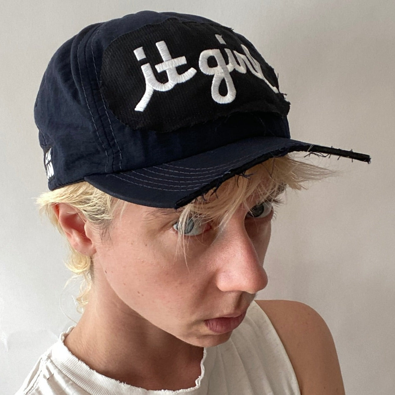 model wearing a navy blue baseball cap with copped brim and a black patch sewn over the front with white embroidered cursive reading it girl