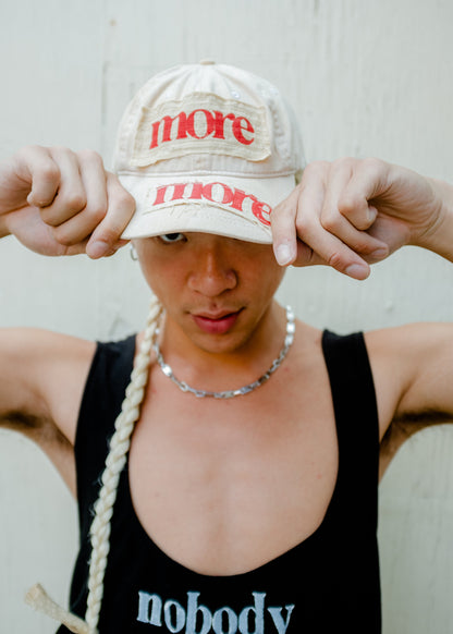 model wearing beige hat with patched with stamped red letters reading more more on the brim and front. hat has blonde extension braid coming from the back to mimic a rattail 