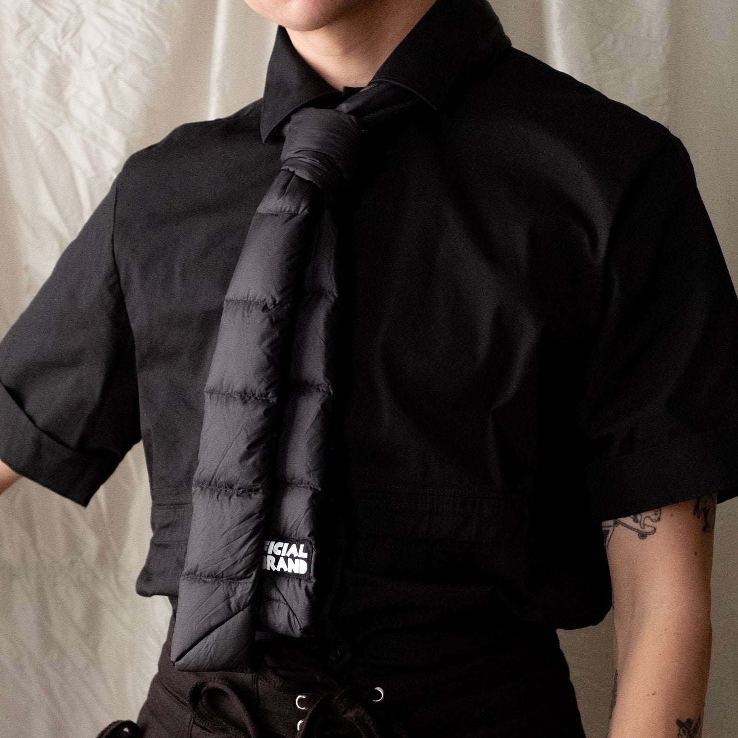 black puffer tie tied on model wearing a black short sleeved button down. Back of tie is visible to show tag reading OFFICIAL REBRAND