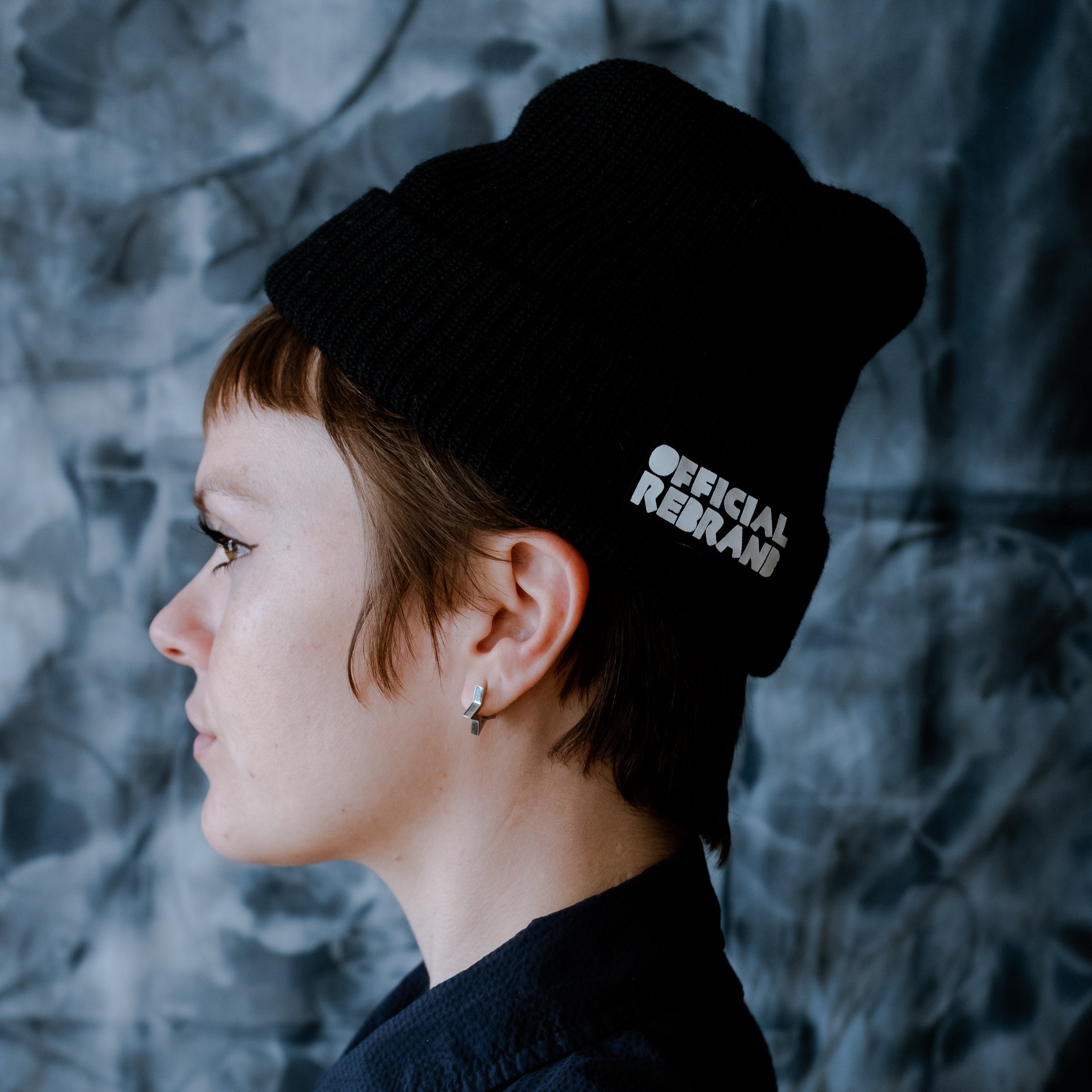 profile of model wearing black wool beanie with white text reading Official Rebrand on the tag