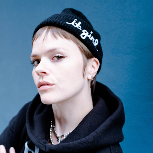 model wearing black wool beanie with white cursive embroidery reading it girl