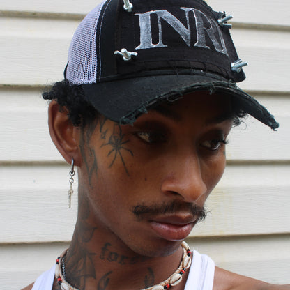 model wearing chopped brim trucker hat with white mesh back, black patch with white INRI stamp, and nuts and bolts affixing the four corners of the patch to the front of the hat