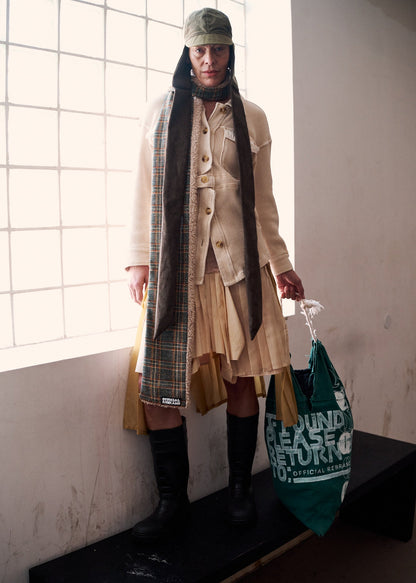 Person wearing a peach pleated collage skirt, beige coat and plaid scarf with a green bag, standing against a white wall with a bright window.
