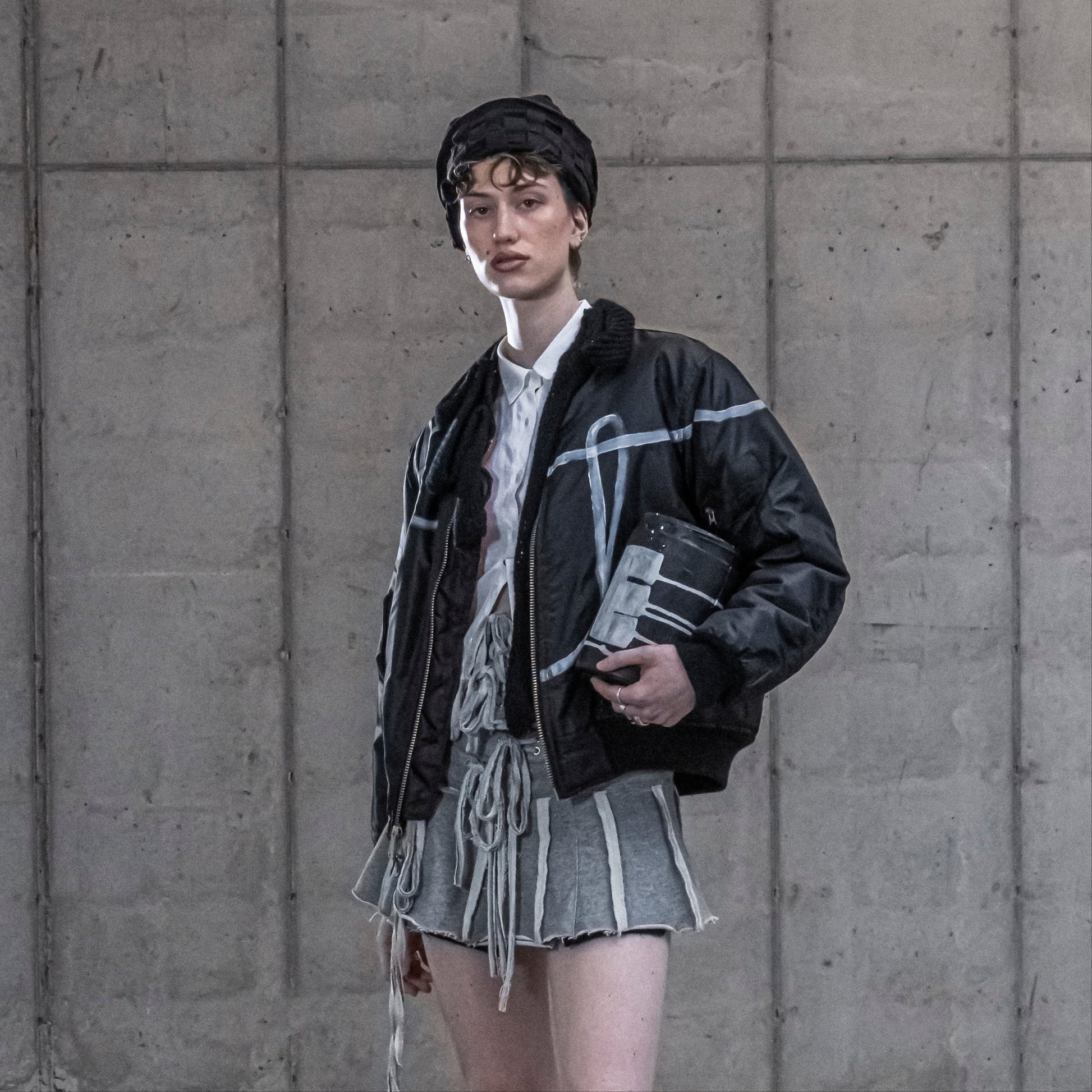 Person wearing a black jacket and gray skirt against a concrete wall