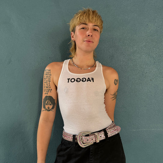 model standing wearing a white a-line tank top, ribbed, with black screen printed text reading TOGGAF. model is in front of a teal wall