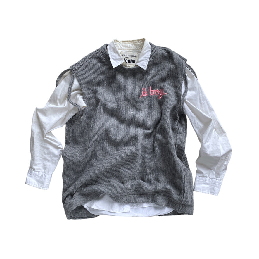 gray knit sweater vest with slits around the arm holes and pink cursive embroidery on the chest reading it boy posed with a white button down underneath 