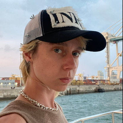 model posing in front of waterway wearing trucker hat with white mesh back, white patch with black INRI stamp, and nuts and bolts affixing the four corners of the patch to the front of the hat and wrapping around in a crown of thorns 
