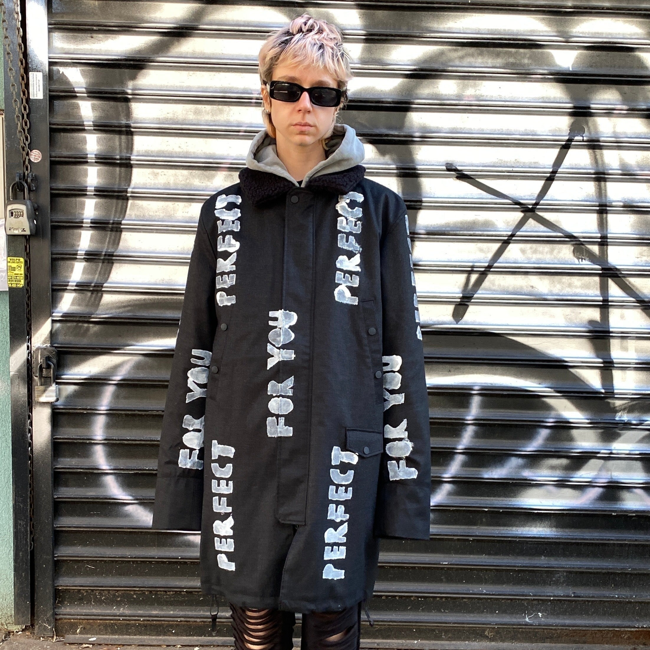 model wearing sunglasses and black long winter coat with faux shearling collar and white hand-painted stenciled PERFECT FOR YOU text across the front and arms 