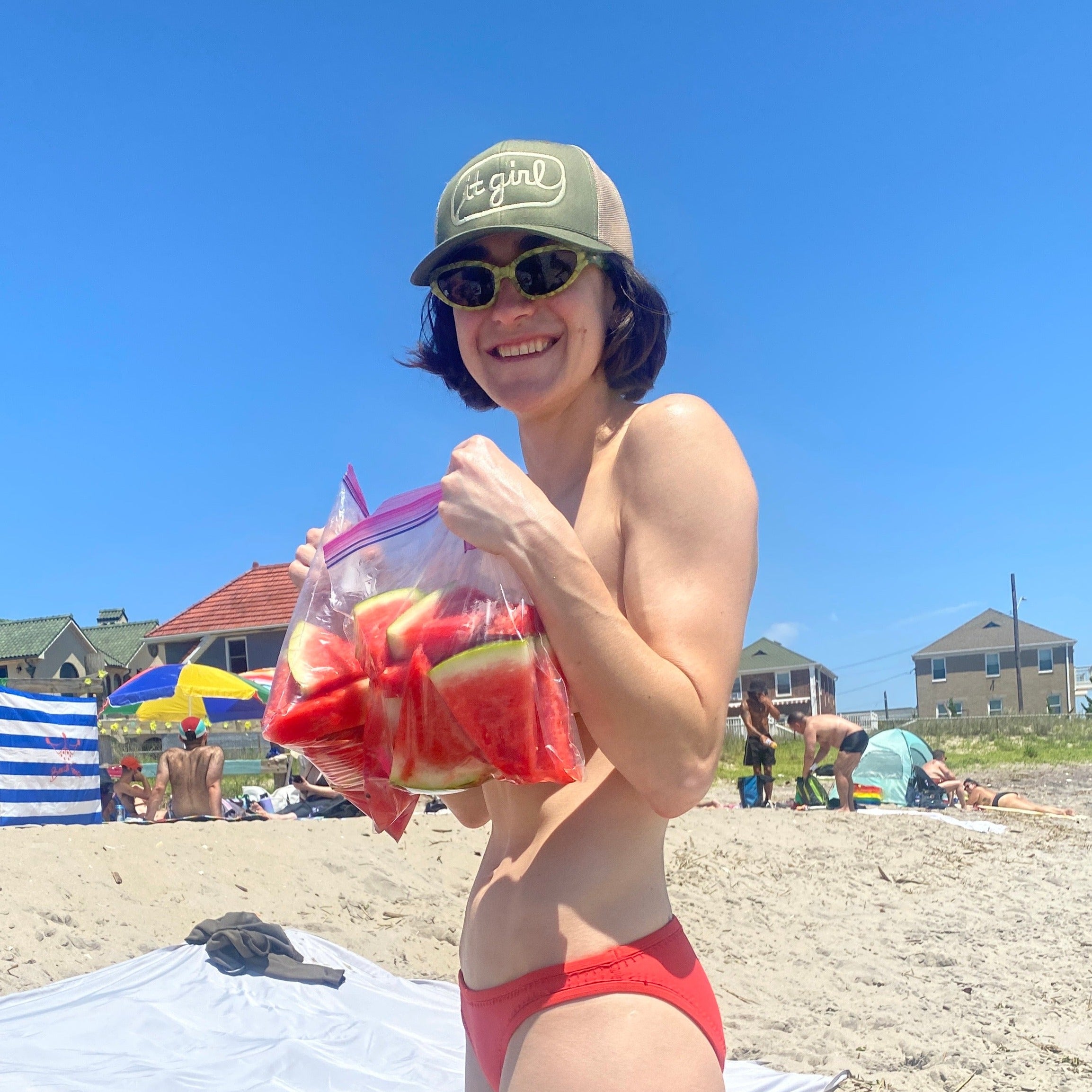 model at the beach holding watermelon wearing olive green and beige khaki trucker hat with khaki embroidery reading it girl in cursive