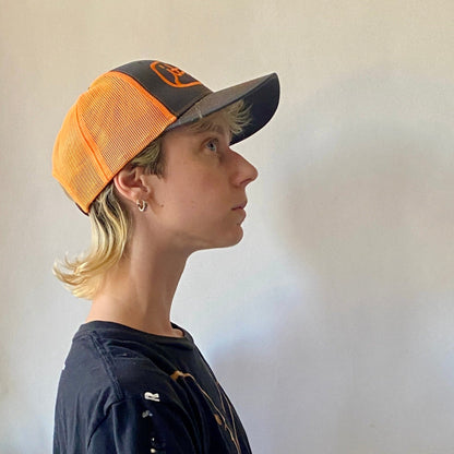 profile of model wearing neon orange and stone gray trucker hat with orange embroidery reading it they in cursive