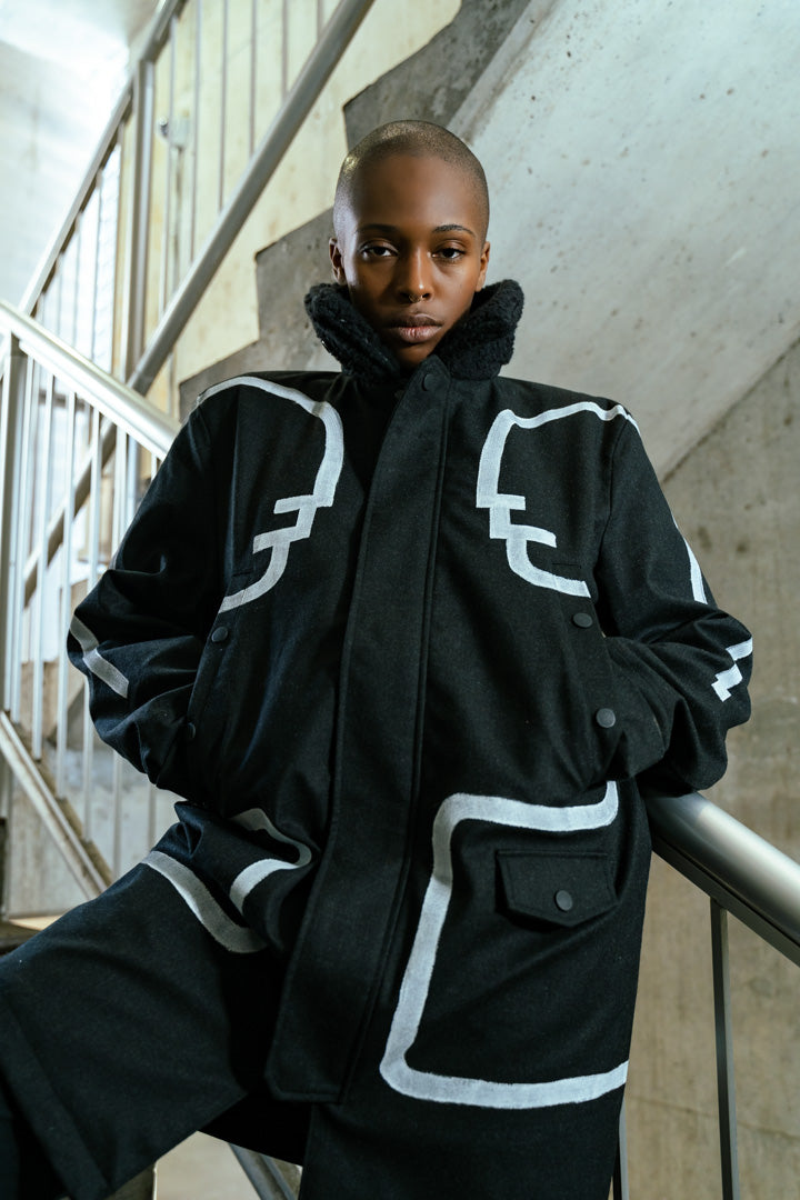 model wearing black long winter coat with faux shearling collar and white conversation figures hand painted to face each other across the winter coat's front and arms