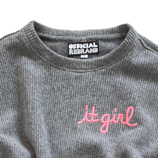 close up of neck and upper chest of gray knit sweater vest showing pink embroidery reading it girl and an inner black tag with white text reading OFFICIAL REBRAND